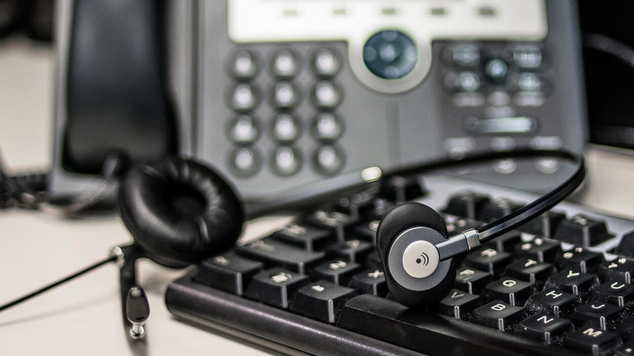 Un casque d’écoute pour téléphone repose sur un clavier d’ordinateur. À l’arrière-plan, on aperçoit un téléphone à touches.
