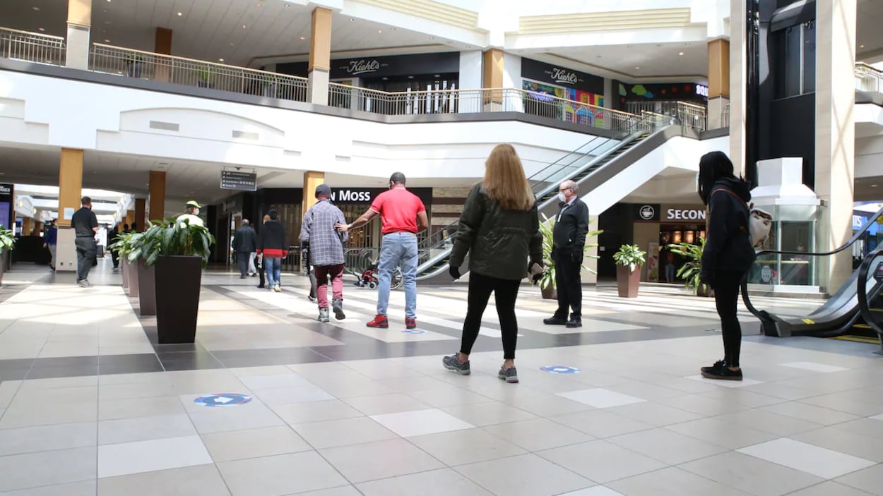 Des gens de dos debout dans un centre commercial. Un employé, qui porte un masque, les surveille pour s'assurer qu'ils respectent les règles de distanciation physique.