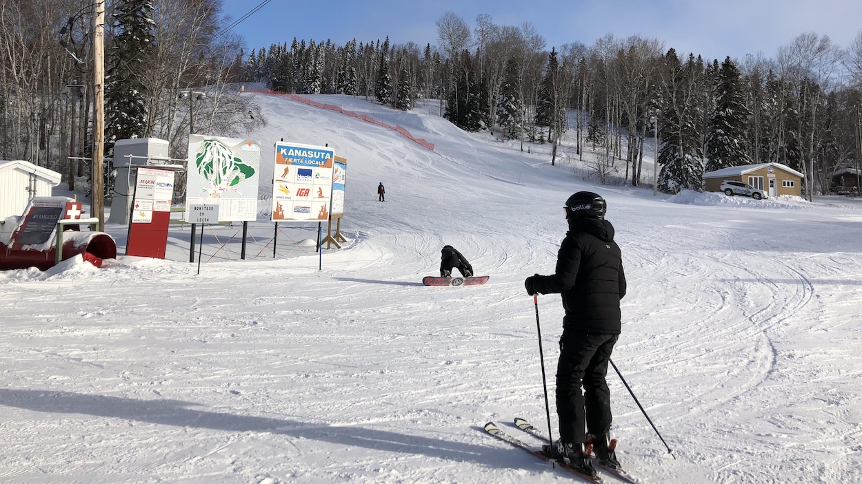 Un skieur attend au pied du Mont Kanasuta, pendant qu'un planchiste s'apprête à terminer sa descente.