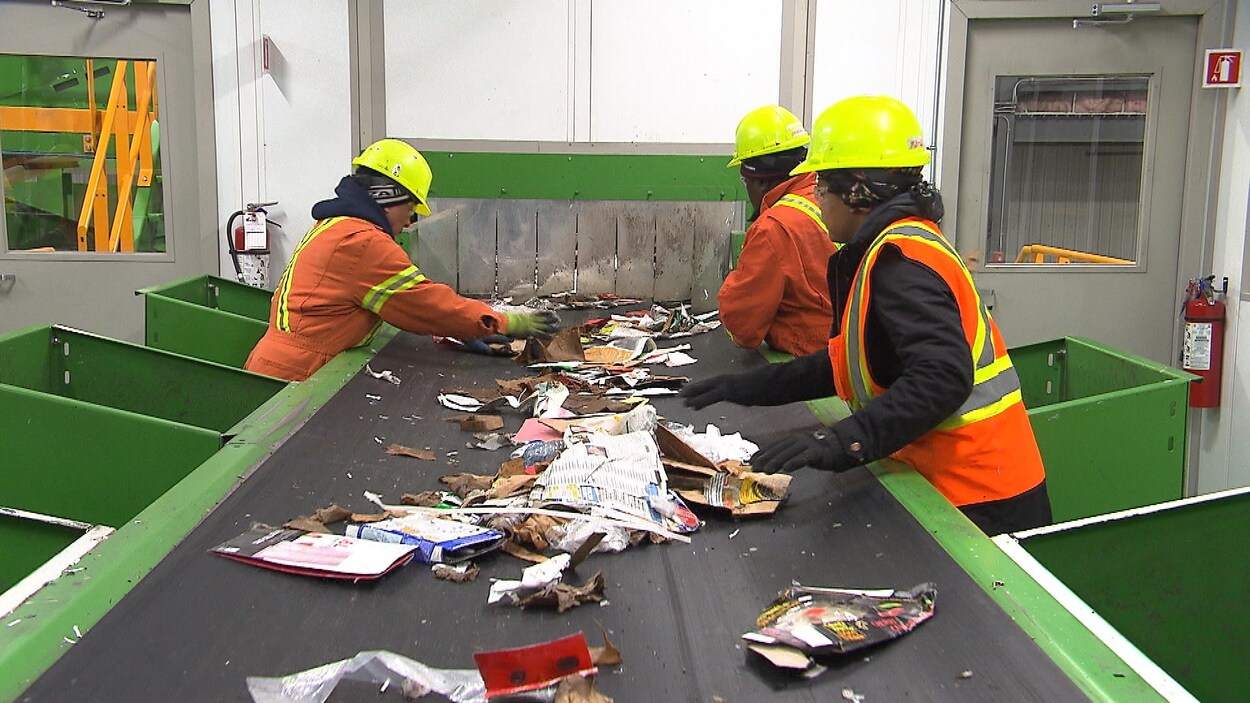 Des gens vêtus de casques et de dossards se tiennent autour d'un tapis roulant où défilent des matières recyclables à trier.