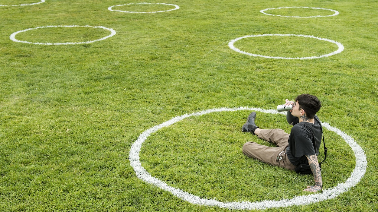 Un homme boit dans une canette, assis seul dans un cercle de distanciation peint dans l'herbe.