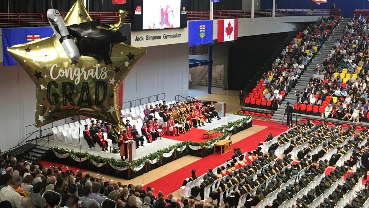 Un ballon indiquant en anglais "félicitations diplômés" flotte dans les airs dans une salle où se déroule une cérémonie de remise de diplômes.