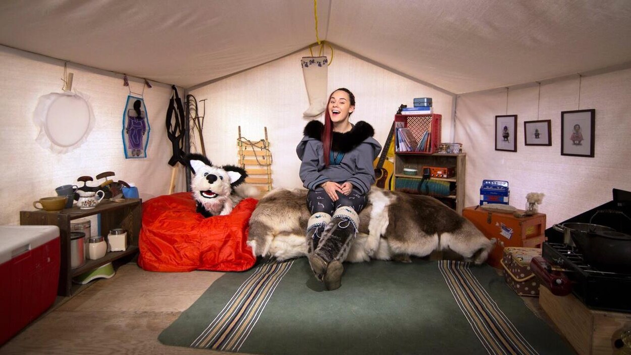 Une femme est assise sur un lit, dans une tente, entourée d'objets faisant référence à la culture inuit.