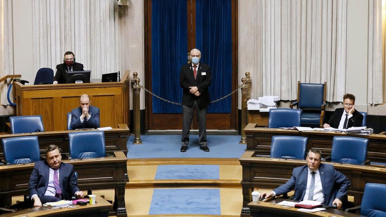 Six hommes dans la Chambre de l'Assemblée législative, dont deux avec des masques, tous à deux mètres les uns des autres.