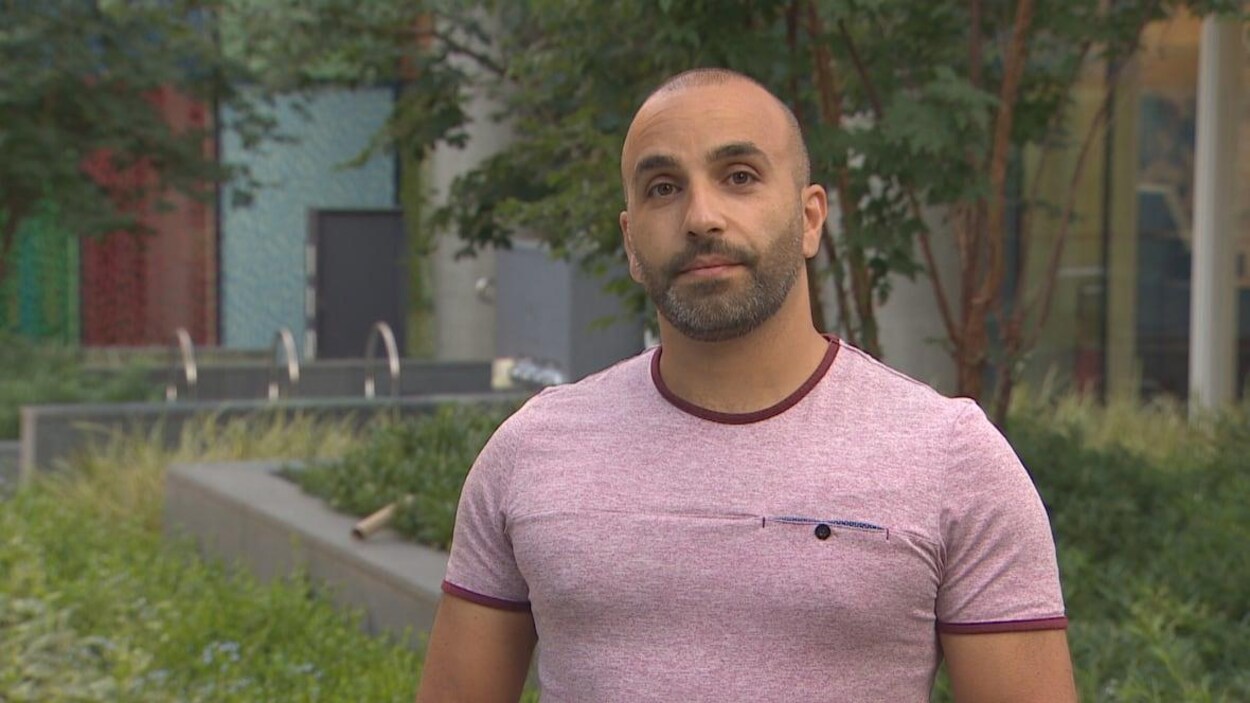 Un homme regarde devant. Il porte un t-shirt et se trouve dans un décor urbain.