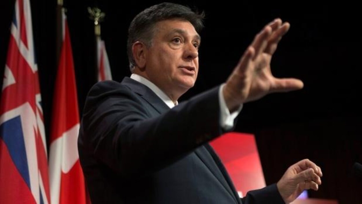 Charles Sousa qui parle devant des drapeaux dans une salle à Queen's Park.
