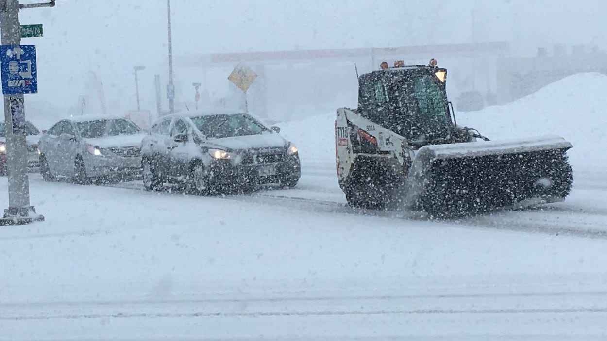 Un chasse-neige dégage une rue, suivi d'une file de voitures.