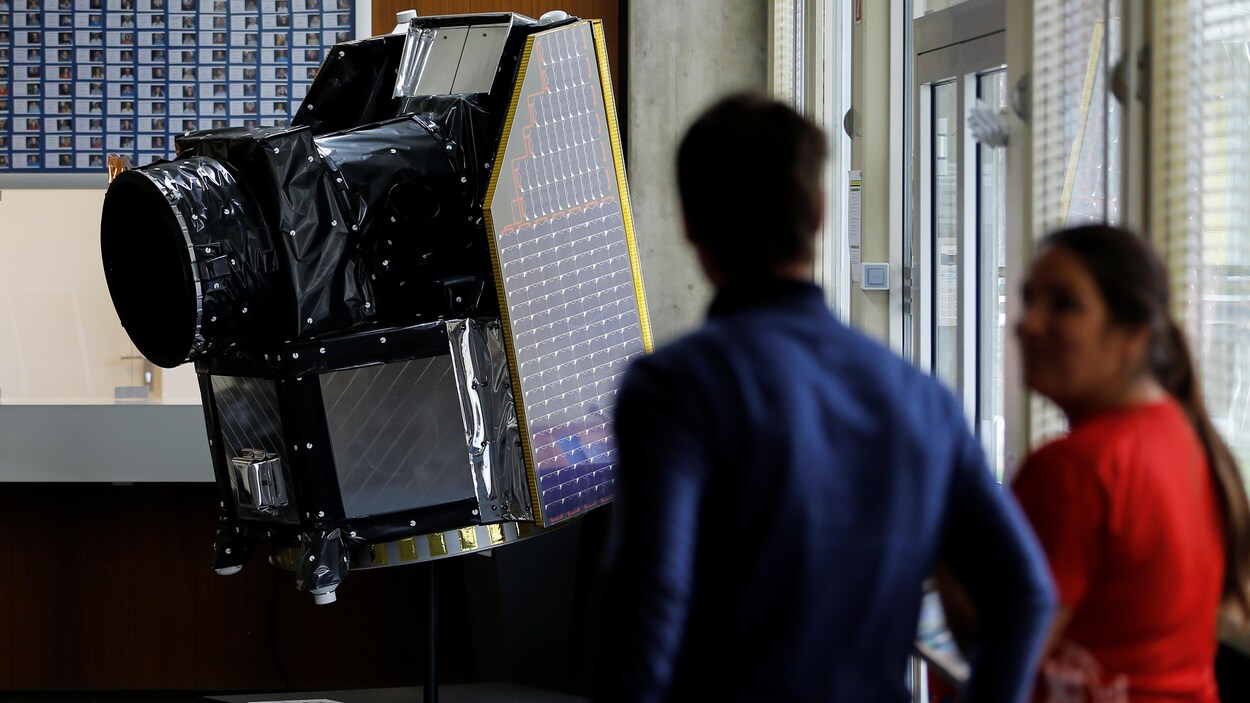 Deux personnes discutent à proximité du téléscope, qui a la forme rectangulaire des premiers appareils photos, mais en bien plus gros et avec des panneaux solaires sur le côté.