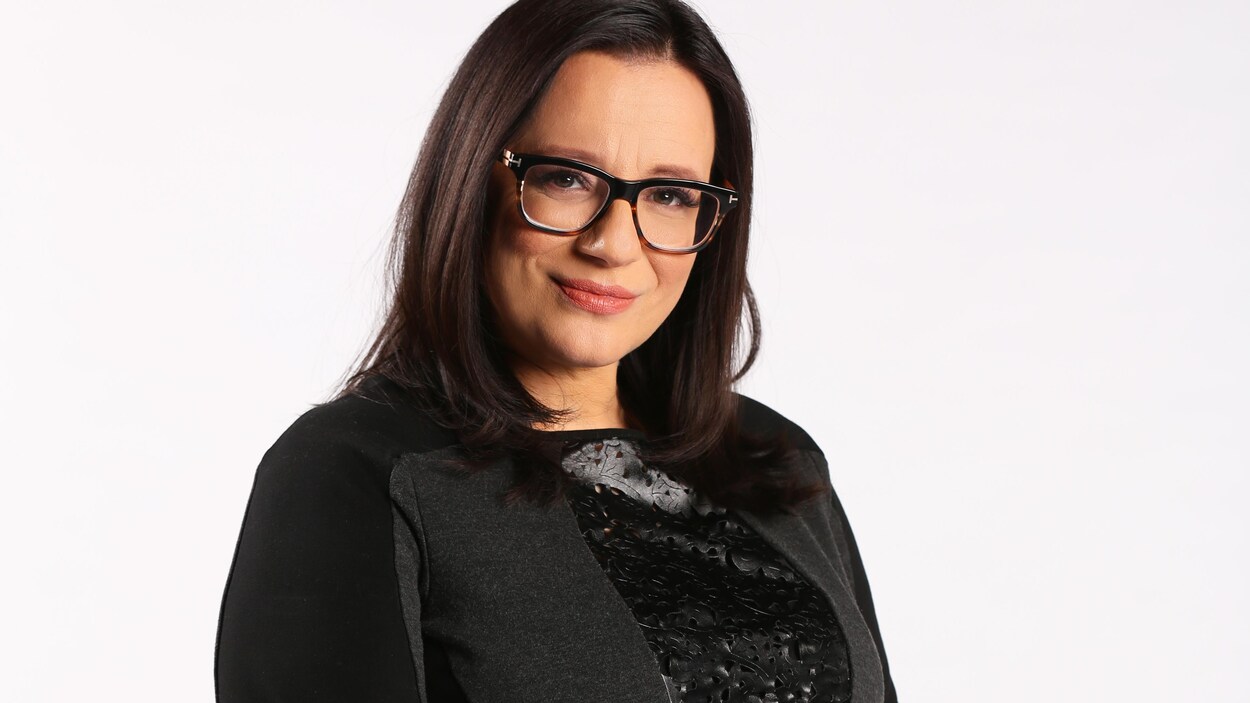 Portrait en couleur de l'autrice autochtone Cherie Dimaline, sur fond blanc. Elle a les cheveux lâchés, porte des lunettes, un chandail noir à manches trois-quarts, et a des tatouages sur les avant-bras.