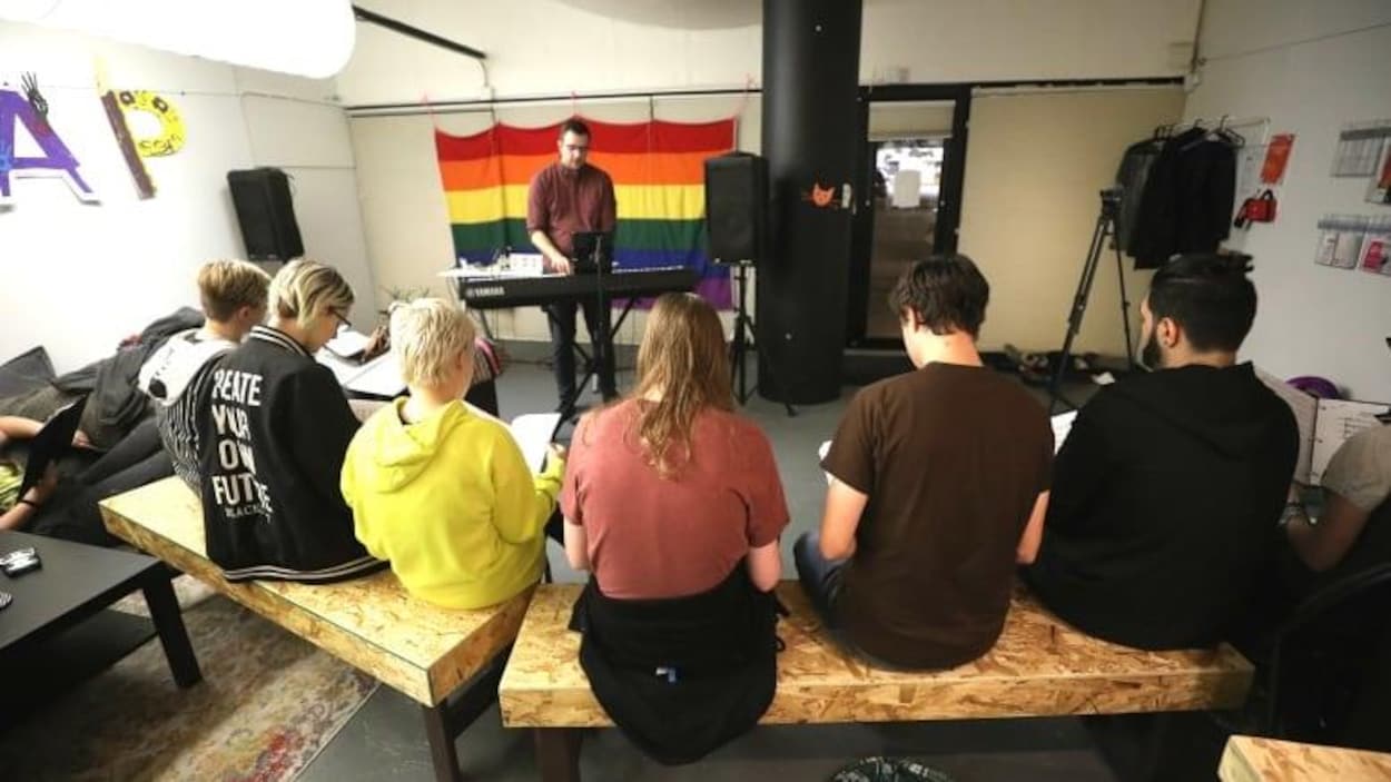 Une personne joue au piano dans une salle. Des jeunes sont assis sur un banc et chantent.