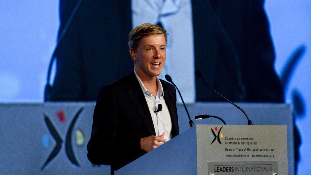 Une photo de Chris Hughes en train de donner un discours devant un écran géant rediffusant son image.