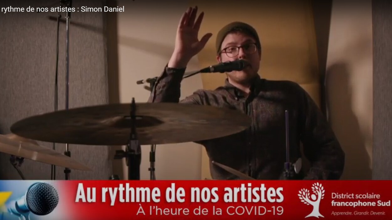 Simon Daniel assis à sa batterie, saluant la caméra.