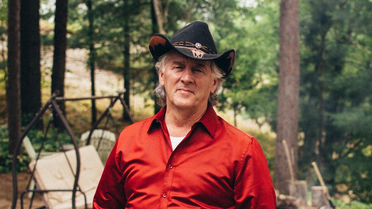Un homme est debout dans la forêt. Il porte un chapeau et une chemise rouge.