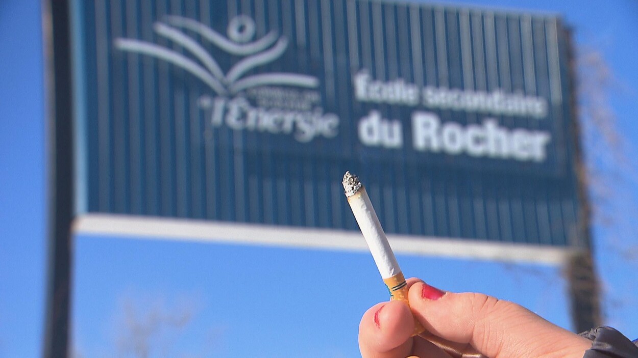 Une femme tient une cigarette devant une affiche d'école.