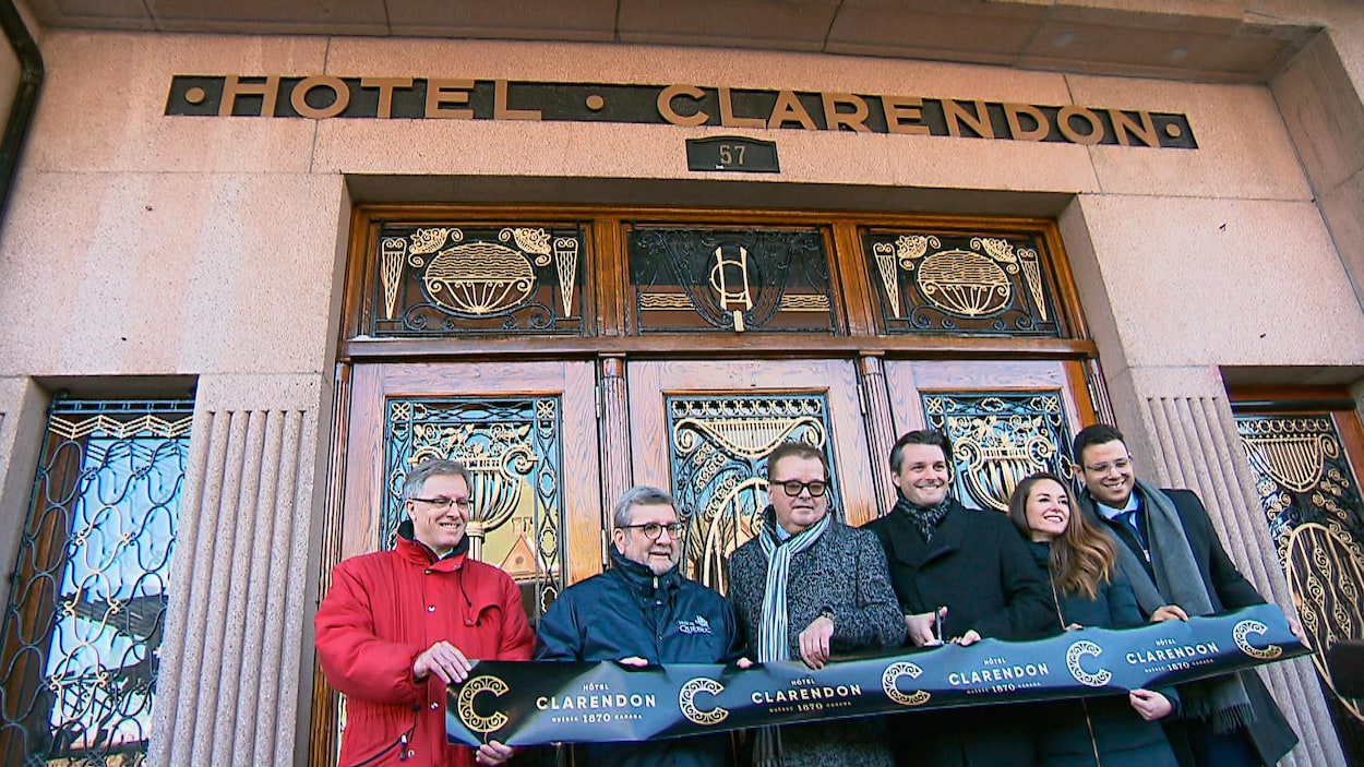 Six personnes souriantes tiennent une banderole sur laquelle il est écrit Hôtel Clarendon. Une des personnes tient aussi des ciseaux et coupe la banderole.