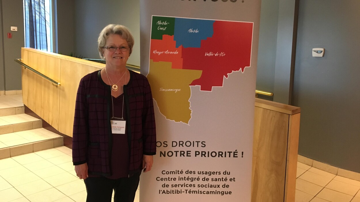 Une femme pose devant une affiche du Comité des usagers du Centre intégré de santé et de services sociaux de l'Abitibi-Témiscamingue.