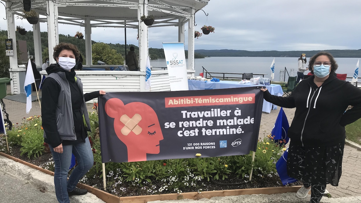 Deux femmes tiennent une affiche indiquant « Abitibi-Témiscamingue. Travailler à se rendre malade, c'est terminé. 131 000 raisons d'unir nos forces ».