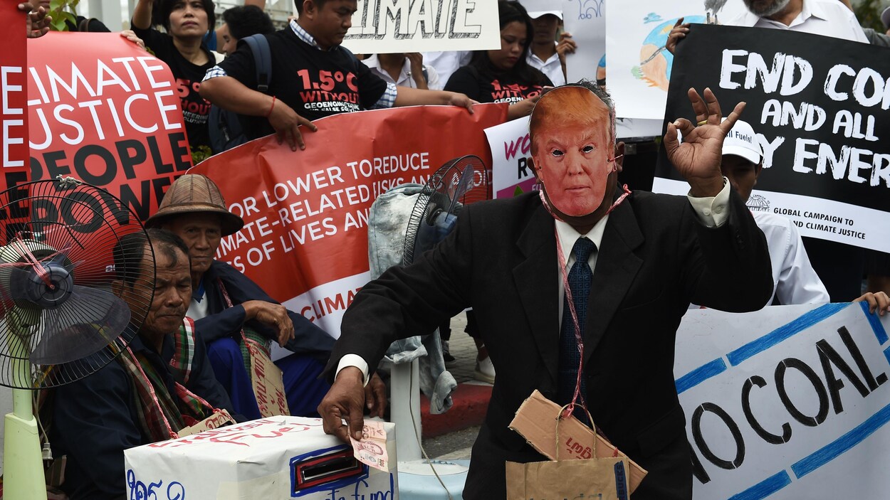 Un homme déguisé en Donald Trump gesticule devant plusieurs manifestants brandissant diverses pancartes.