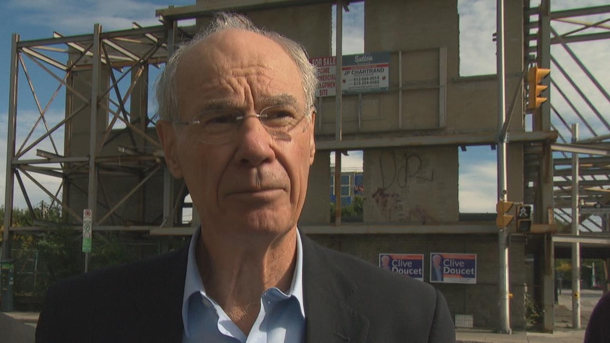 Clive Doucet en point de presse devant un bâtiment désaffecté.