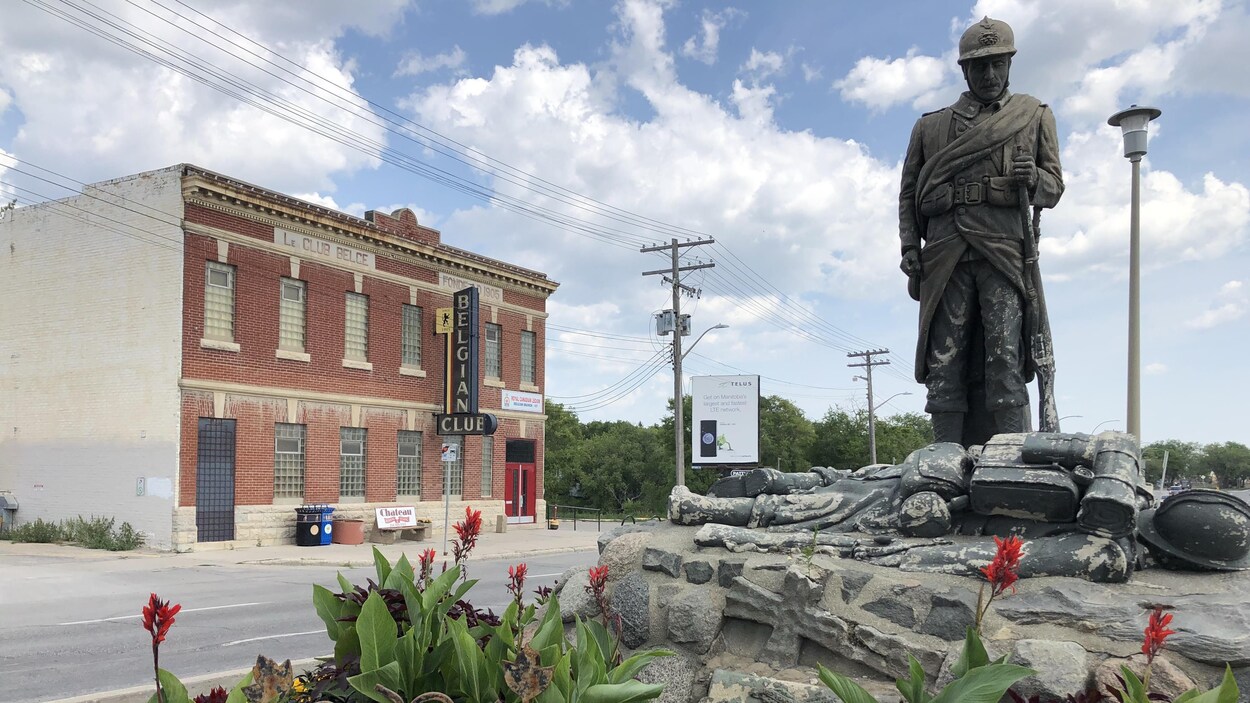 Une statue représentant des combattants de la Première Guerre mondiale se trouve sur le terre-plein devant un bâtiment en brique rouge avec une enseigne où il est écrit « Belgian Club ».