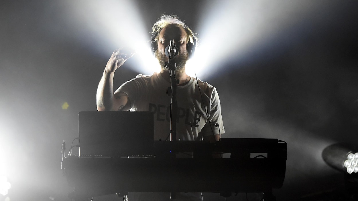 Un musicien portant des écouteurs se tient derrière un ordinateur portable et une table de mixage, sur une scène de spectacle.