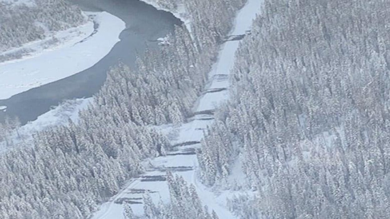 Une route enneigée près d'une rivière, des dizaines d'arbres sont couchés au travers de la chaussée.