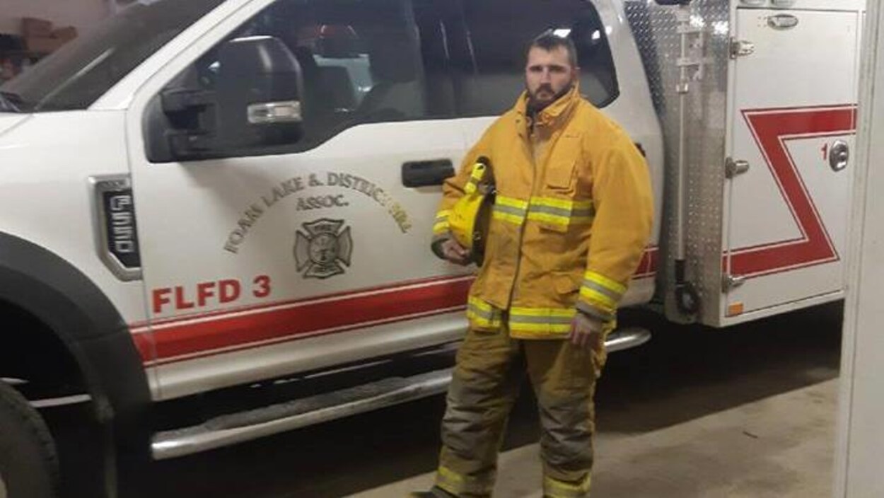 Le pompier bénévole Cole Maksymytz tient son casque sous le bras devant une ambulance.