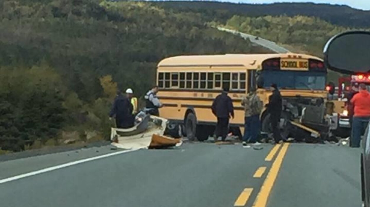 Un policier et des témoins entourent un autobus scolaire à l'avant endommagé et des débris d'un véhicule accidenté sur une route