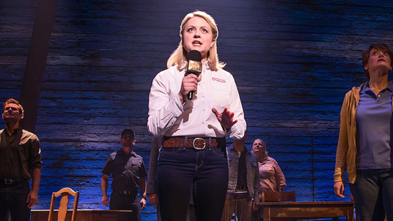 Une scène de la comédie musicale « Come From Away », où l'on peut voir une actrice jouant une journaliste, entourée d'autres acteurs.