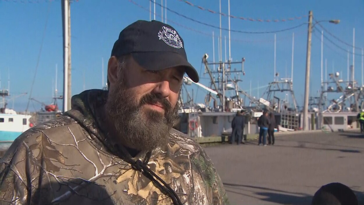 Joël Comeau interviewé sur un quai devant des bateaux de pêche.
