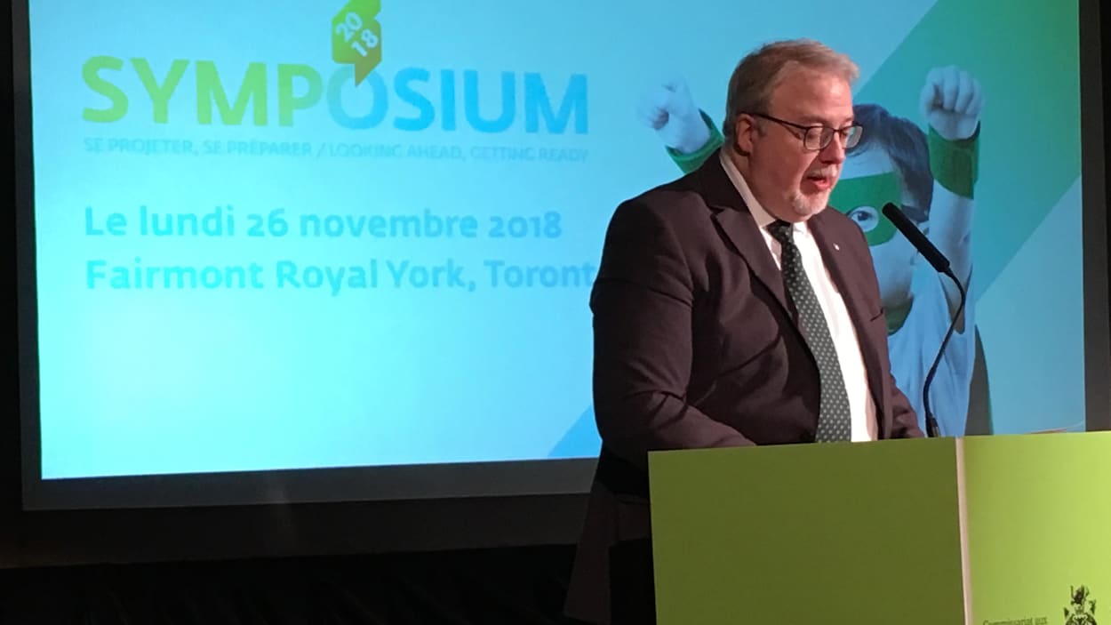 Photo d'un homme en complet au podium; on peut lire le mot symposium sur un écran derrière lui.