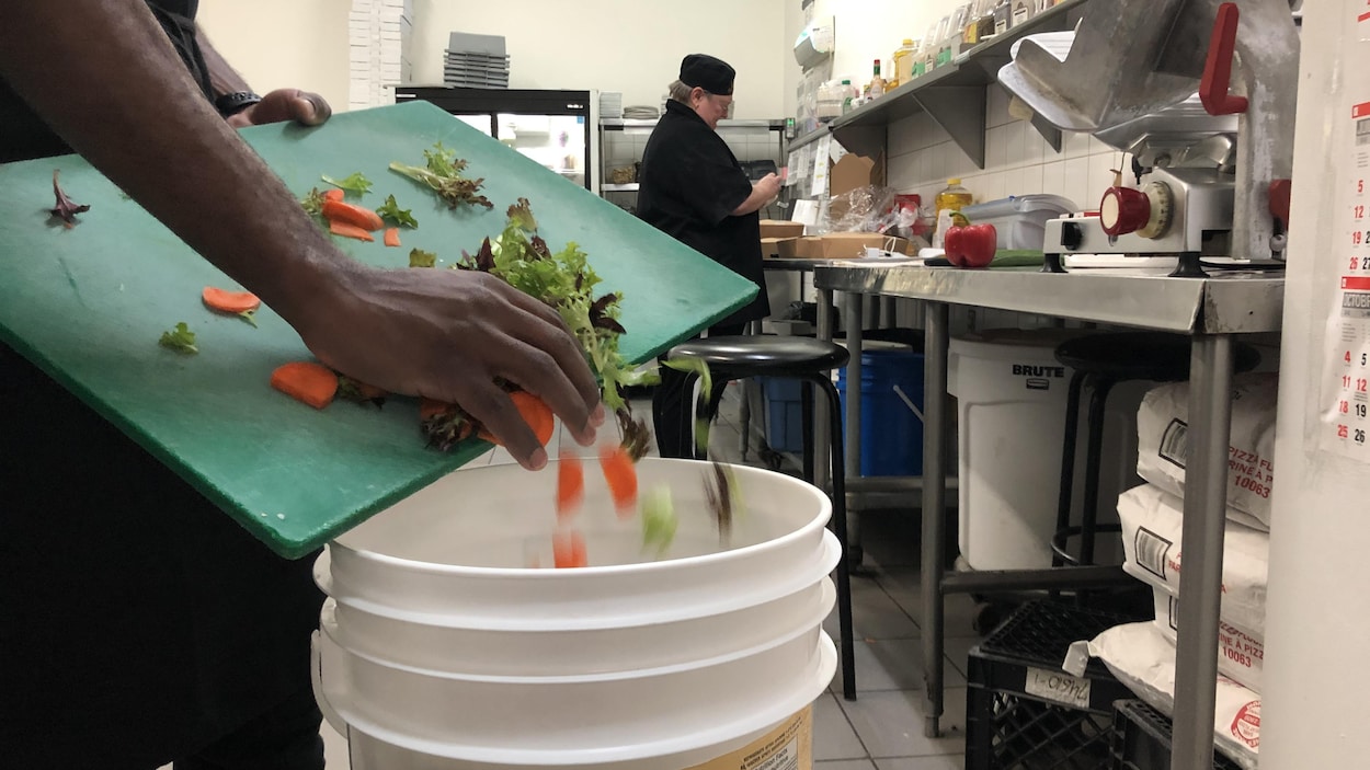 Une main pousse des retailles de légumes d'une planche à découper dans une chaudière, dans une cuisine de restaurant.