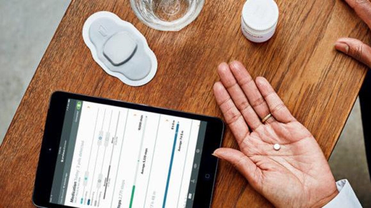 Une photo prise du dessus dans laquelle on peut voir la main d'une personne tenant un petit comprimé blanc et une table sur laquelle sont déposés un iPad, un timbre médical, un verre d'eau et un flacon de médicaments.