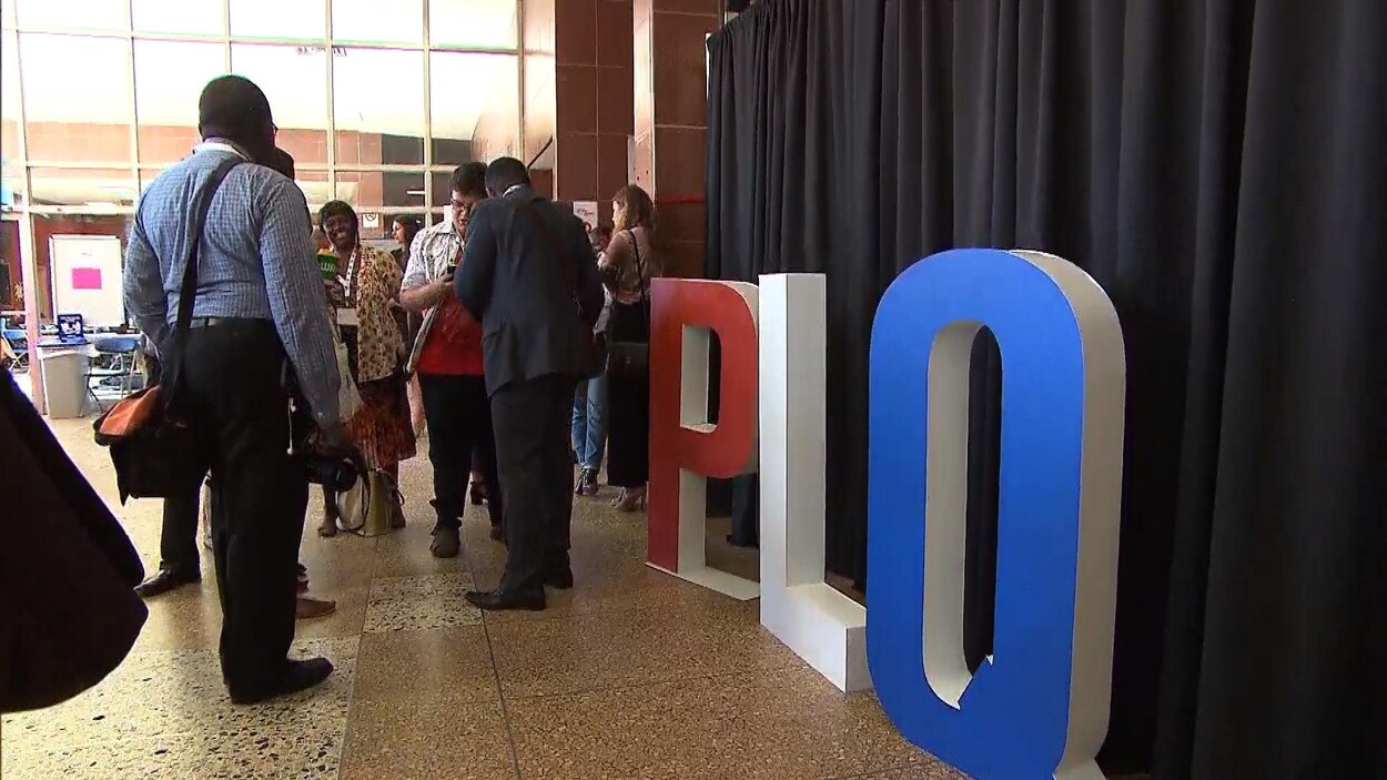Plusieurs personnes entrain de discuter dans une salle à côté d'une installation à l'effigie du PLQ Plusieurs personnes entrain de discuter dans une salle à côté d'une installation à l'effigie du PLQ