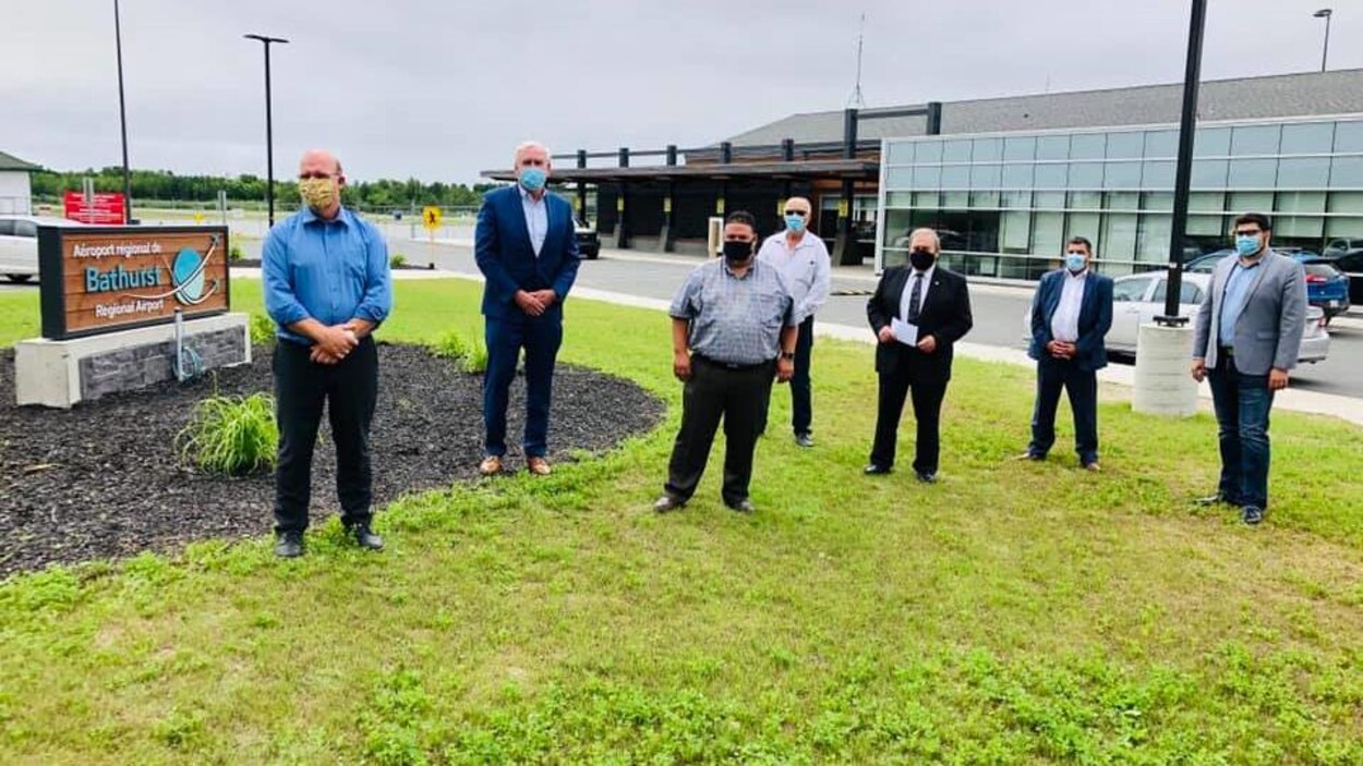 7 hommes portant un masque de protection se tienne debout à l'extérieur devant l'aéroport régional de Bathurst.