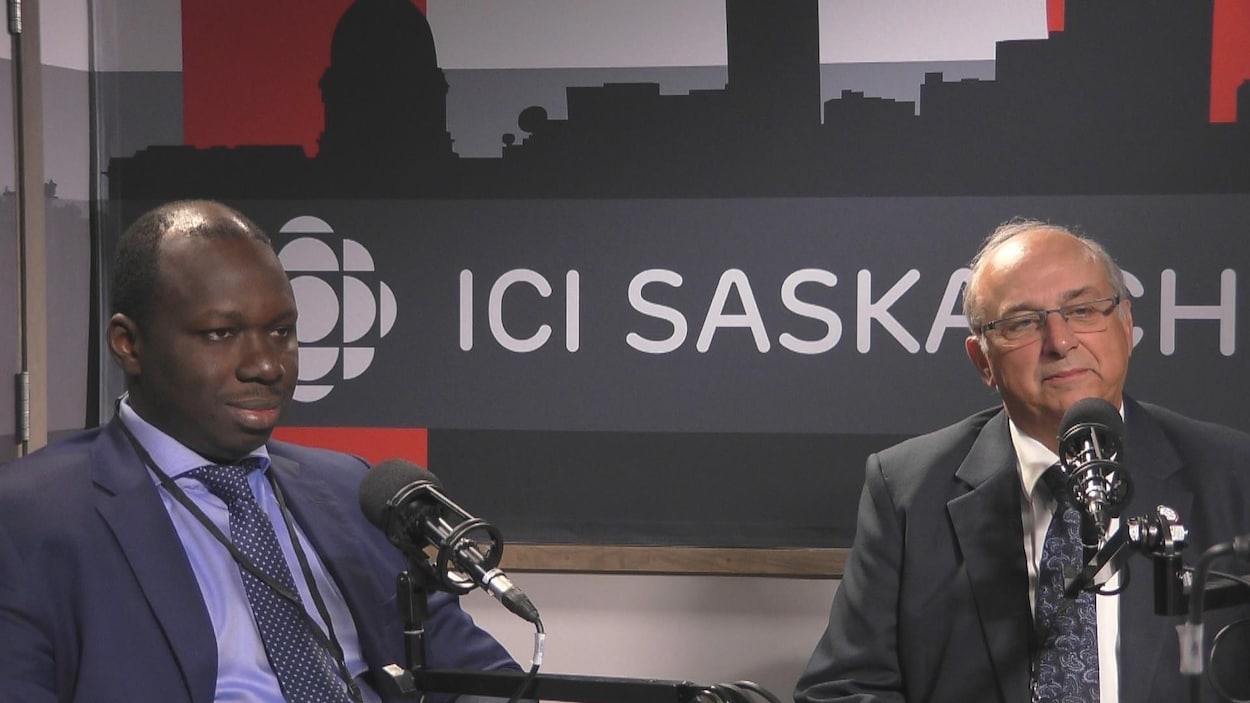 Deux hommes, en costume cravate, assis derrière des micros avec en arrière-plan une affiche d'ICI Saskatchewan.