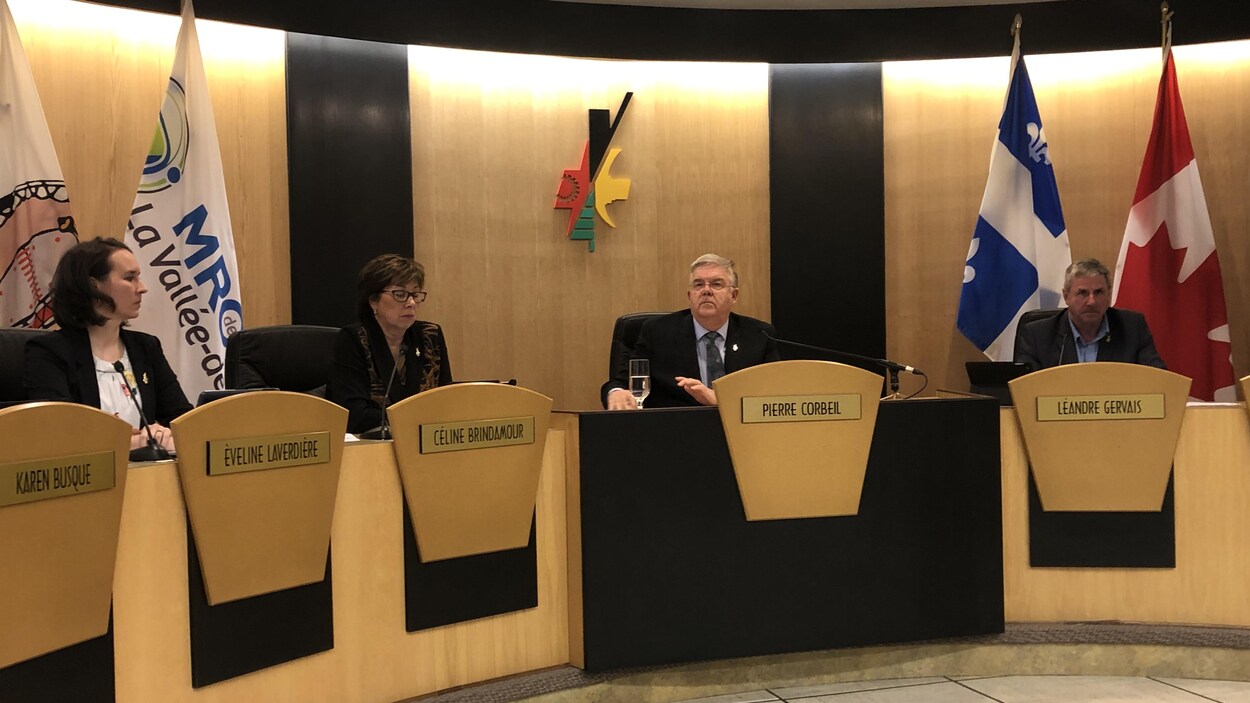 Trois élus et le maire Pierre Corbeil assis à leur place assignée dans la salle du conseil municipal.