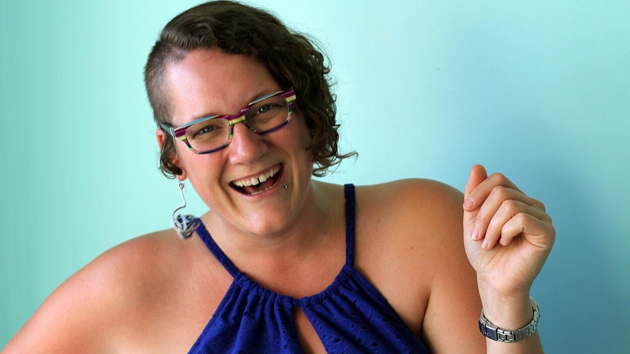 Une femme très souriante a des cheveux bruns rasés d'un côté et frisés au menton de l'autre côté. Elle porte des lunettes avec une monture multicolore et une robe sans manches bleue.
