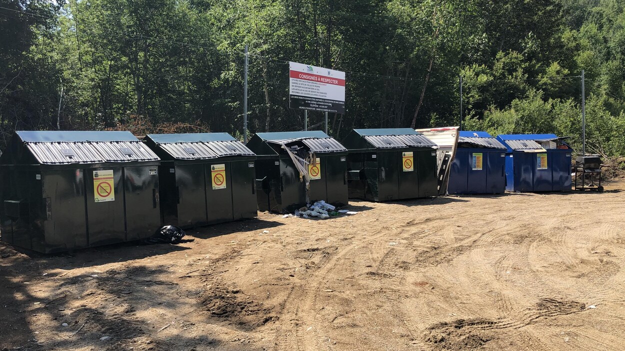 Six gros conteneurs à déchets sont alignés au fond d'un terrain sablonneux.