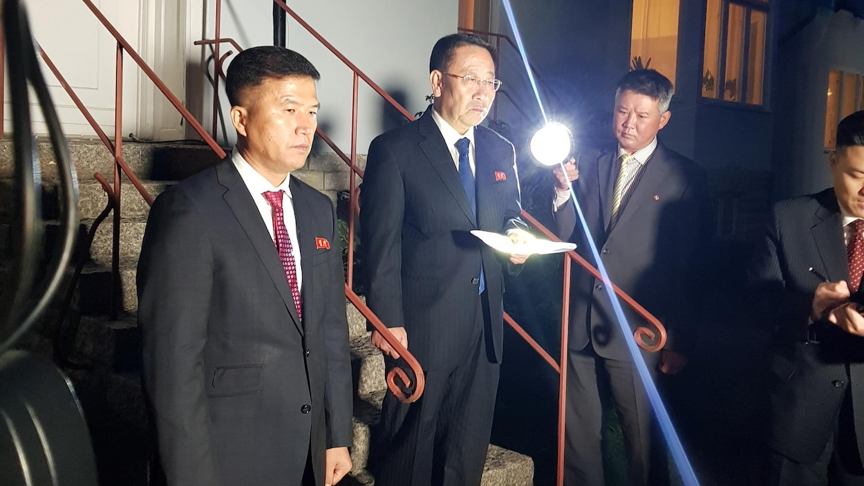 Kim Myong Gil, feuilles à la main dans un escalier, entouré de trois hommes et éclairé par une forte lumière.