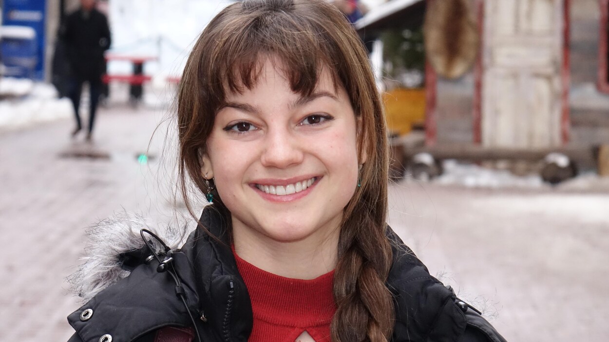 Une jeune femme sourit à la caméra, ses cheveux bruns noués en une longue tresse sur le côté gauche.
