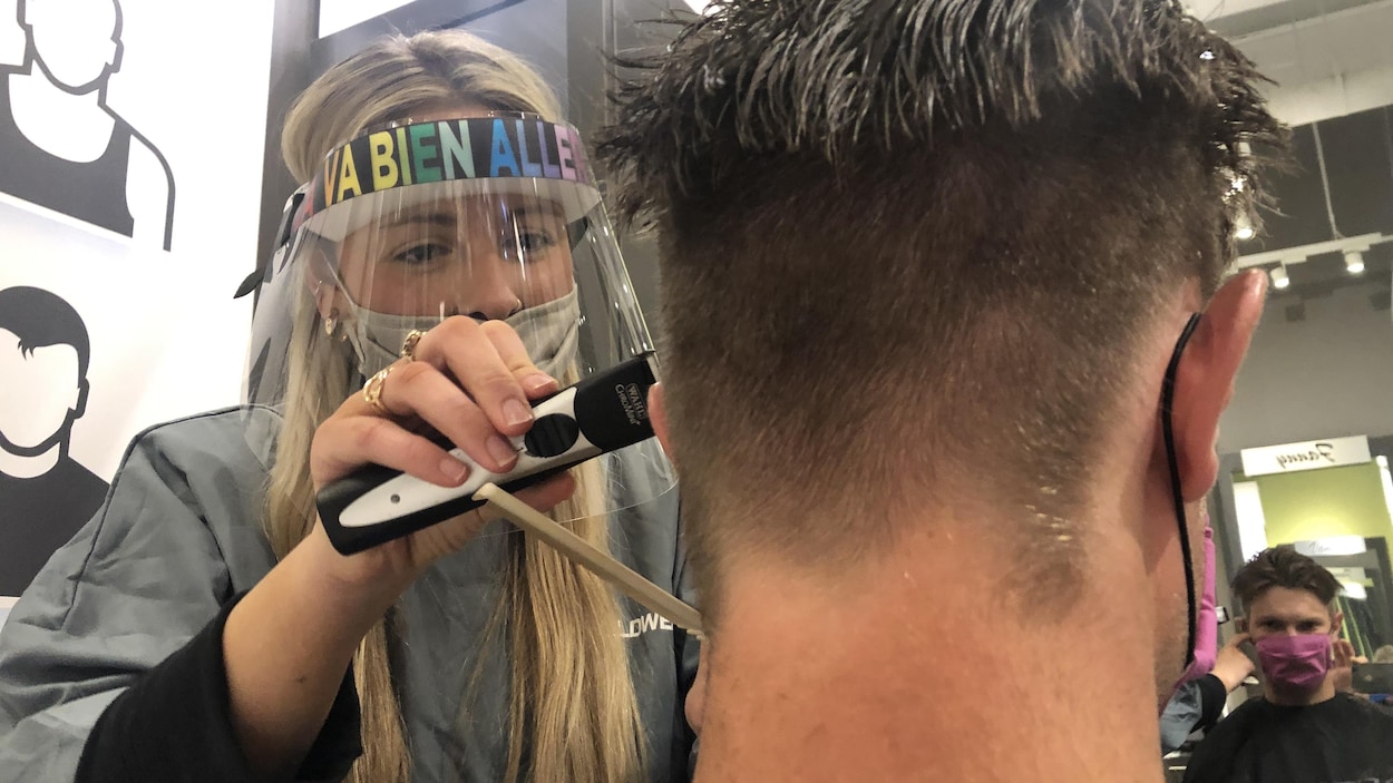 Une coiffeuse, portant un équipement de protection contre le coronavirus, rase les cheveux d'un client.