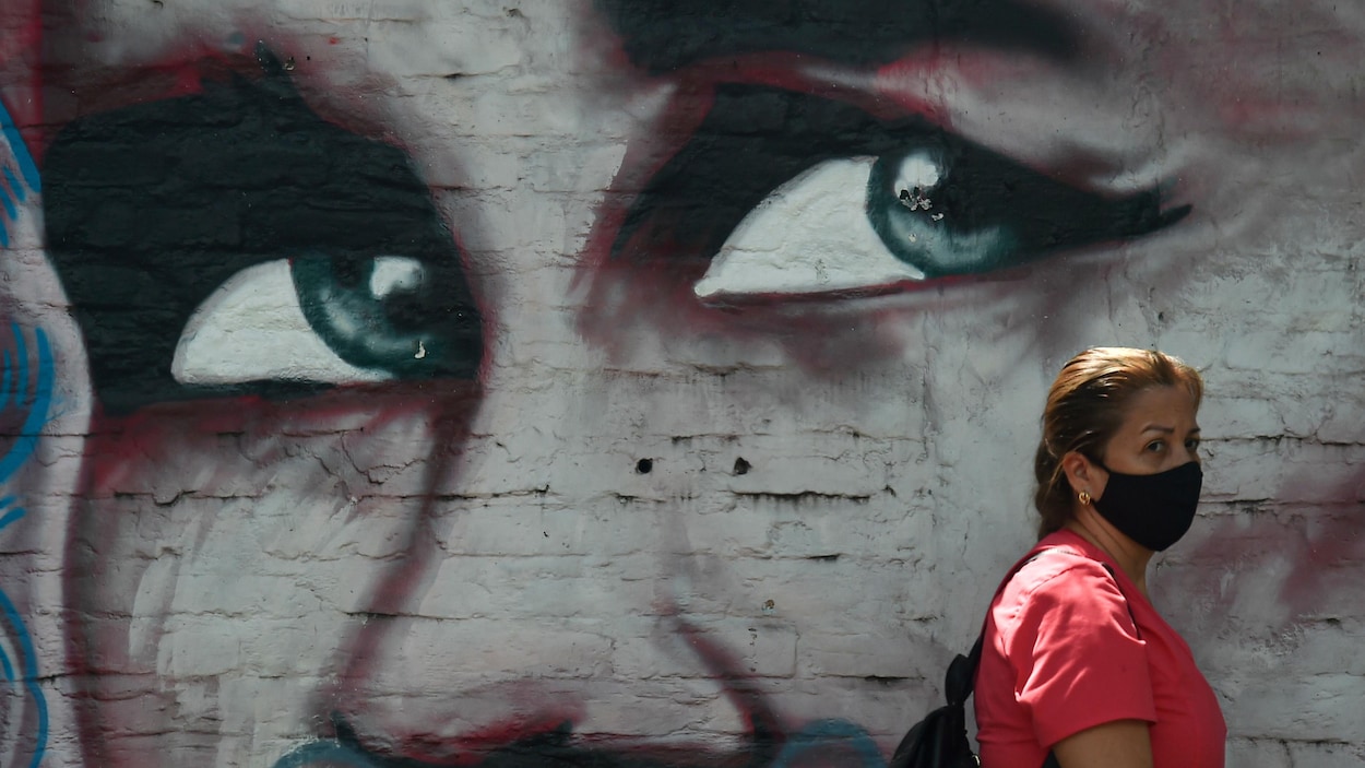 Une femme masque passe devant un graffiti dépeignant, en gros plan, le visage inquiet d'une femme.