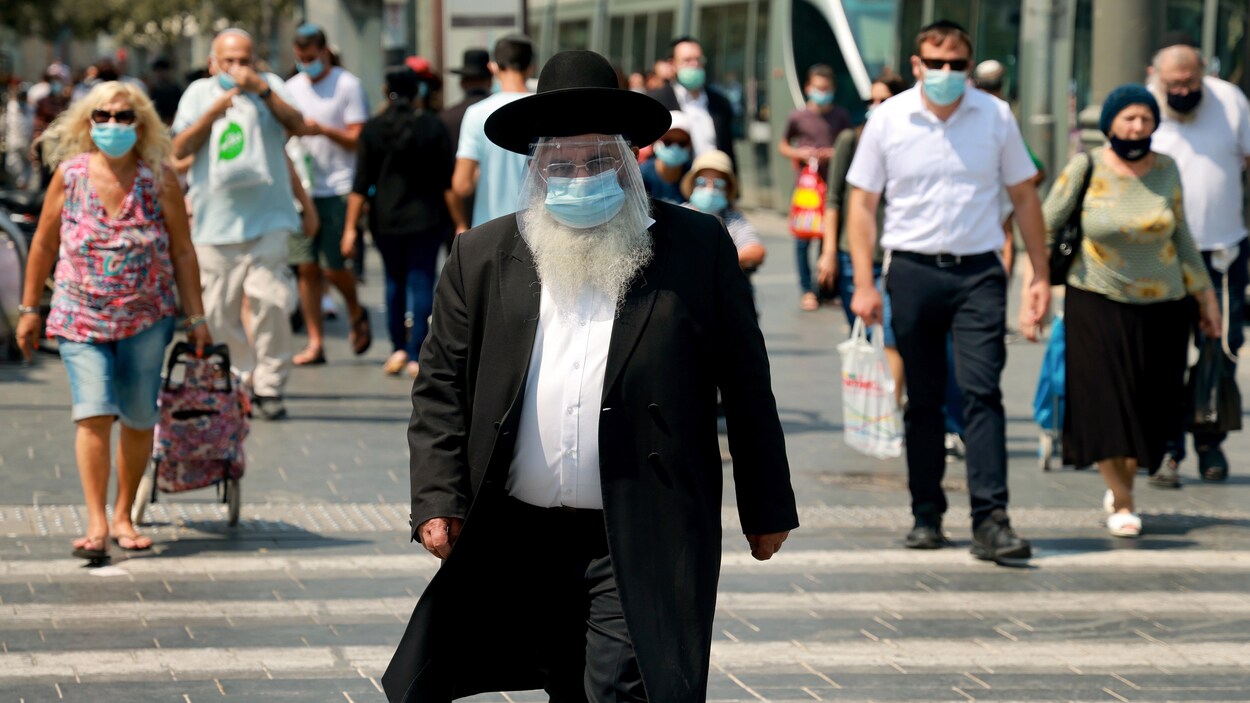 Un homme juif ultra-orthodoxe portant un masque et une visière marche dans la rue. Des passants portent le masque.