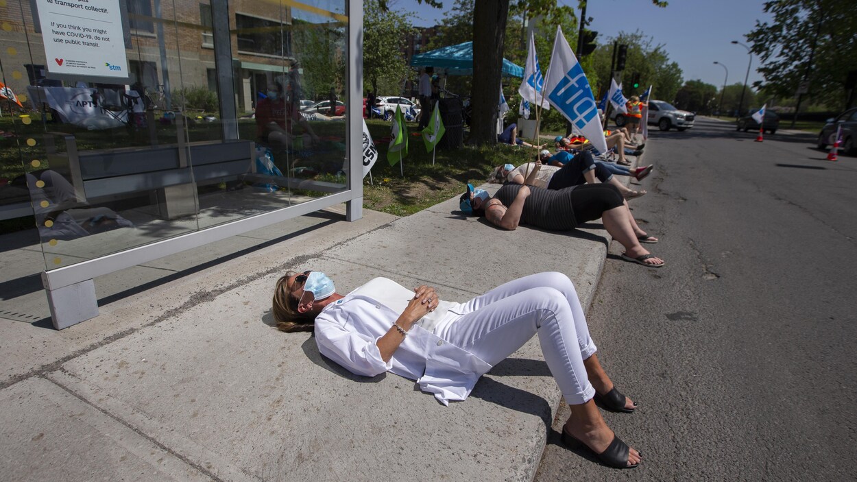 Du personnel infirmier est couché sur le trottoir de manière symbolique lors d'une manifestation devant un établissement, en plein jour.