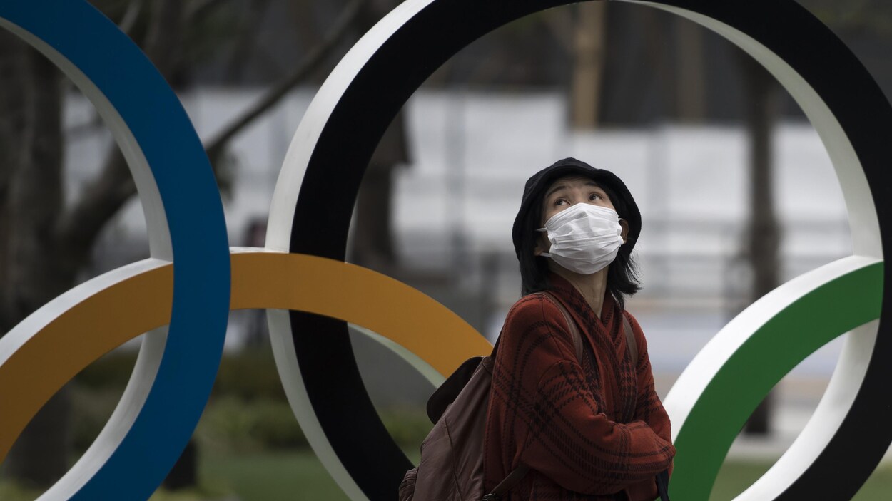 Une femme avec un masque passe devant les anneaux olympiques.