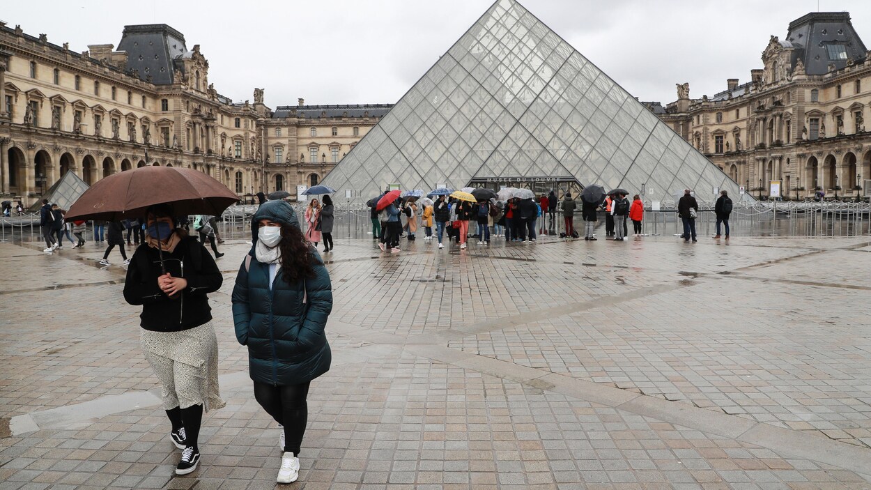 Deux femmes masquées marchent près de la pyramide du Louvre.