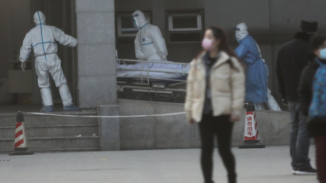 Des employés portant un uniforme de protection les couvrant complètement font rouler une civière à l'extérieur de l'hôpital.