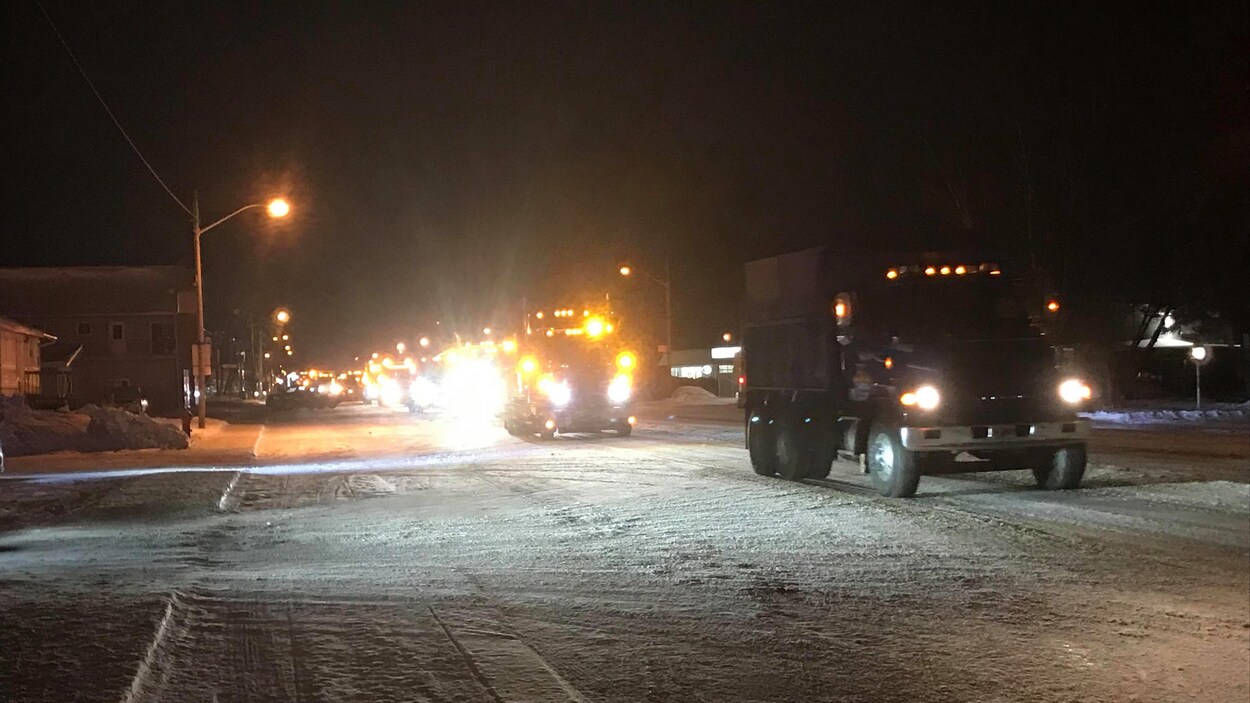 Des camions circulent sur une rue enneigée, en soirée.