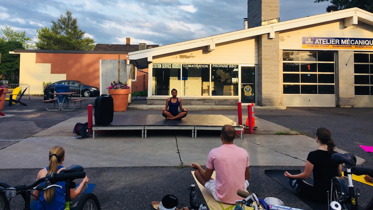 Une femme sur une plateforme enseigne le yoga en plein-air, devant la station-service désaffectée.